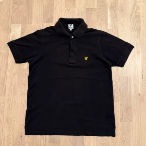 Svart pikétröja från Lyle & Scott - Säljer en klassisk svart pikétröja från Lyle & Scott med korta ärmar och en gul broderad logotyp på bröstet. Perfekt för en stilren och avslappnad look. Tröjan har en krage med knappar för en snygg finish. Pris kan diskuteras.