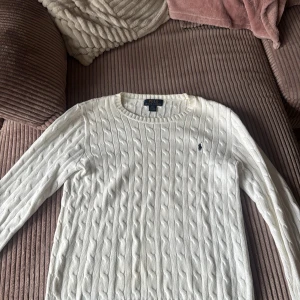 Vit stickad tröja från Ralph Lauren - Säljer en stilren vit stickad tröja från Ralph Lauren med kabelstickat mönster och det klassiska logotypbroderiet på bröstet. Tröjan har långa ärmar och rund halsringning. Perfekt för en elegant och avslappnad look. Den e storlek L men passar även mig som har S 