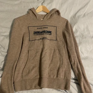 Beige hoodie från Zadig & Voltaire - Säljer en snygg beige hoodie från Zadig & Voltaire med tryck på framsidan. Tröjan har en klassisk känguruficka och ribbade muddar vid ärmslut och nederkant. Perfekt för en avslappnad stil.