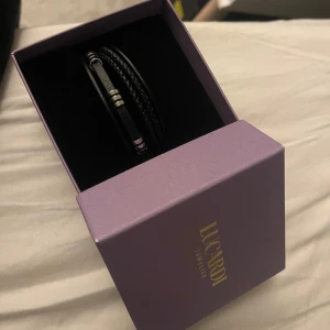 Svart armband från Lucardi - Stilrent svart armband från Lucardi med flätade detaljer och silverfärgade accenter. Perfekt för att ge en modern touch till din stil. Kommer i en elegant lila presentask. Oanvänd då den inte riktigt passar klädstilen jag personligen har, men väldigt fin och i perfekt kvalite