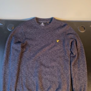 Lyle & Scott hoodie  - Säljer en stilren grå tröja från Lyle & Scott med deras ikoniska gula logga på bröstet. Tröjan är långärmad och har en klassisk rund halsringning. Perfekt för en avslappnad och snygg look. Helt ny.