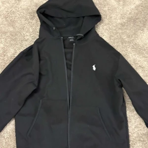 Svart huvtröja från Polo Ralph Lauren - Säljer en stilren svart huvtröja från Polo Ralph Lauren med dragkedja och broderad logga på bröstet. Tröjan har långa ärmar och en praktisk huva. Perfekt för en avslappnad look. Pris går att diskutera ✅✅