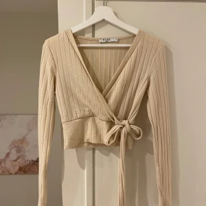 Beige ribbad omlottopp från NA-KD - Säljer en stilren beige ribbad omlottopp från NA-KD. Toppen har långa ärmar och knytning i midjan för en snygg passform. Perfekt för både vardag och fest!