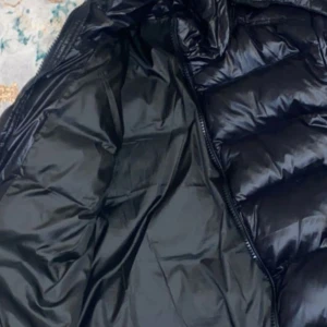 Svart pufferjacka med huva - Säljer en svart pufferjacka med huva. Jackan har en glansig yta och är utrustad med dragkedja framtill. Perfekt för kyligare dagar med sin vadderade design.