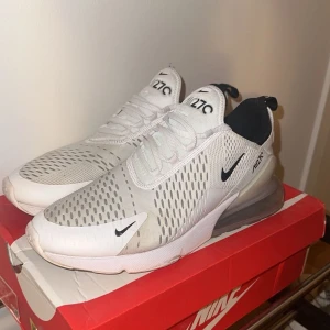 Nike Air Max 270 i vitt och svart - Snygga Nike Air Max 270 sneakers i vitt med svarta detaljer. Skorna har en luftig design med perforerade partier och en stor synlig Air-enhet i hälen för extra komfort. Perfekta för en sportig look. Adda snap adidasbyxor om du är intresserad!