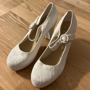 Vita pumps med spetsdetaljer från Vox - Eleganta vita pumps från Vox med vacker spetsdetalj och en dekorativ knapp på sidan. Skorna har en rund tå och en stabil klack, perfekt för speciella tillfällen.
