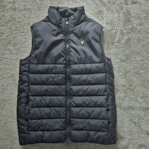 Svart quiltad väst från Lyle & Scott - Snygg svart quiltad väst från Lyle & Scott med dragkedja och ett litet gult emblem på bröstet. Perfekt för lager-på-lager under kyliga dagar. Passar ungdomar i åldern 14-15 år.