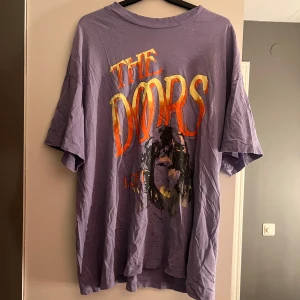 Lila t-shirt med The Doors-tryck - Cool lila t-shirt med stort The Doors-tryck i orange och gult på framsidan. Perfekt för fans av bandet! T-shirten har en avslappnad passform och korta ärmar. Oversize i storlek, använd 1 gång 