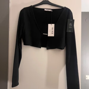 Svart croppad kofta från NA-KD - Säljer en stilren svart croppad kofta från NA-KD. Koftan har långa ärmar och en enkel design som passar perfekt till olika outfits. Den är i storlek M och har en modern look.