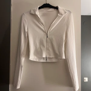 Vit ribbad croppad tröja med dragkedja - Snygg vit ribbad tröja med dragkedja framtill. Tröjan är croppad och har långa ärmar, perfekt för en stilren look. Passar bra till både jeans och kjol.