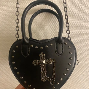 Svart hjärtformad handväska med kedja - Snygg svart hjärtformad handväska med nitar och ett dekorativt kors framtill. Väskan har en kedja som axelrem och två handtag för olika bäralternativ. Perfekt för att ge en edgy touch till din outfit.