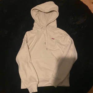 Beige hoodie från Levi's - Säljer en stilren beige hoodie från Levi's med en klassisk känguruficka och justerbar huva. Perfekt för en avslappnad look. Den har en liten logotyp på bröstet. 