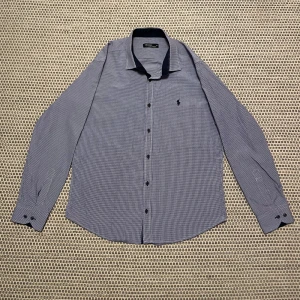 Blå rutig skjorta från Ralph Lauren - Snygg blå rutig skjorta från Ralph Lauren med broderad logga på bröstet. Skjortan har långa ärmar och knappar framtill. Perfekt för en stilren look. Den är inte använd. Storlek XL men passar även om man brukar ha L eller M.