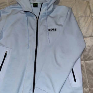 Ljusblå hoodie från Boss - Snygg ljusblå hoodie från Boss med dragkedja och justerbar huva. Den har svarta detaljer vid dragkedjan och fickorna, samt Boss-loggan på bröstet. Perfekt för en avslappnad stil.