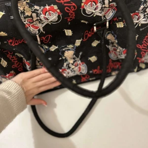 Svart väska med Betty Boop-motiv - Cool svart väska med ett färgglatt Betty Boop-motiv. Väskan har två handtag och är perfekt för att bära med sig det nödvändigaste. Den har ett unikt mönster med text och bilder som verkligen sticker ut.