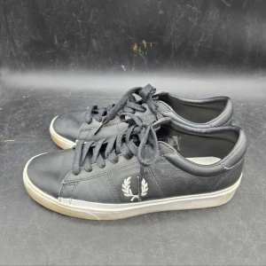 Svarta sneakers från Fred Perry - Snygga svarta sneakers från Fred Perry med vit sula och klassisk logga på sidan. Skorna har snörning och är tillverkade i skinn, vilket ger en stilren look. Perfekta för en avslappnad stil.