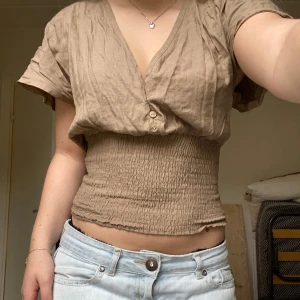 Beige kortärmad topp med smockdetalj - Säljer en beige kortärmad topp med smockdetalj i midjan och v-ringning. Toppen har knappar framtill. Något skrynklig men går att stryka 