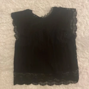Svart spetsblus från ONLY - Snygg svart blus från ONLY med vackra spetsdetaljer. Perfekt för en elegant look. Blusen är ärmlös och har en fin struktur som ger en lyxig känsla. Passar mis som har storlek xs/s. Oanvänd