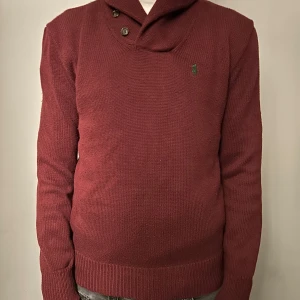 Vinröd stickad tröja från Ralph Lauren - Säljer en stilren vinröd stickad tröja från Ralph Lauren med en liten grön logga på bröstet. Tröjan har en unik krage med knappar och långa ärmar. Perfekt för kyligare dagar!