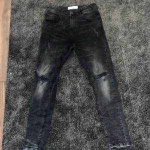 Svarta skinny jeans - Snygga svarta skinny jeans med slitningar på knäna. De har en modern tvättad look och är perfekta för en trendig stil. Midjan är midwaist och storleken är 29/30. Perfekta för att ge din outfit en edgy touch.