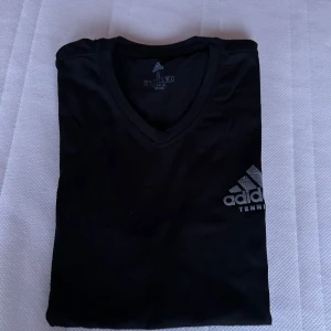 Svart t-shirt från Adidas - Säljer en svart t-shirt från Adidas med v-ringning och korta ärmar. Perfekt för tennis eller träning. Adidas-loggan är tryckt på bröstet. Skön och stilren design.