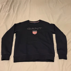 Svart sweatshirt från GANT - Säljer en stilren svart sweatshirt från GANT med broderad låga på bröstet . Tröjan har långa ärmar och en rund halsringning med röd och vit randig detalj på insidan. Skick 8/10 storlek s