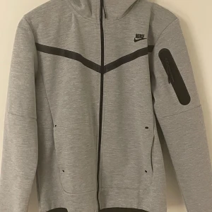 Grå Nike tech fleece och Nike tech byxor  - Passar också som storlek S. Säljer en grå hoodie och matchande byxor från Nike. Hoodien har en dragkedja framtill och en ficka på ärmen. Byxorna har en ficka med dragkedja på sidan. Plagget passar också som storlek S. Perfekt för en sportig look eller avslappnad dag.