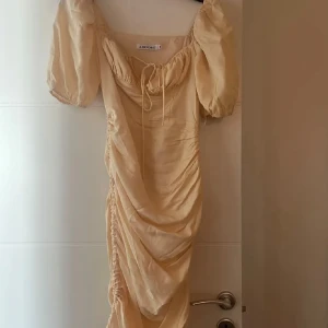 Ny Beige Adoore klänning  - Helt ny Adooreklänning med alla prislappar kvar. Elegant champagnefärgad klänning med puffiga kortärmar och knytning framtill. Klänningen har en smickrande rynkad design längs sidan som ger en unik look. Perfekt för alla tillfällen.