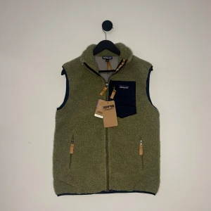 Patagonia Väst - Säljer en olivgrön fleeceväst från Patagonia med dragkedja och en praktisk bröstficka i mörkblått. Västen har en hög krage och detaljer i orange. Perfekt för lager-på-lager under kyliga dagar. Storlek M men passar S. Hör gärna av er vid flera frågor eller funderingar! 