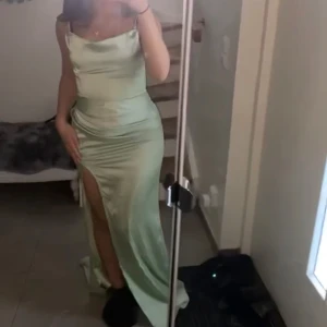 Grön långklänning med slits - Elegant grön långklänning i satin med en stilren slits framtill. Klänningen har en axelbandslös design som ger en sofistikerad look. Perfekt för speciella tillfällen.