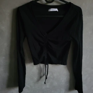 Svart långärmad topp från House Brand - Snygg svart långärmad topp från House Brand med knytning framtill. Perfekt för en stilren look. Tillverkad med miljövänliga material.