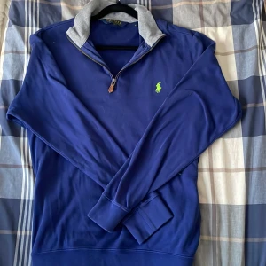 Blå tröja från Polo Ralph Lauren - Säljer en stilren blå tröja från Polo Ralph Lauren med en grå krage och dragkedja. Den har en broderad grön logotyp på bröstet och långa ärmar. Perfekt för en avslappnad och snygg look.