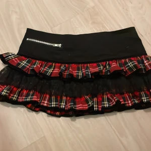 Svart och rutig kjol från Hell Bunny - Snygg kortkjol från Hell Bunny med svart överdel och rutig volang i rött, grönt och gult. Kjolen har en cool dragkedjedetalj och en liten röd rosett. Perfekt för en unik och edgy stil!