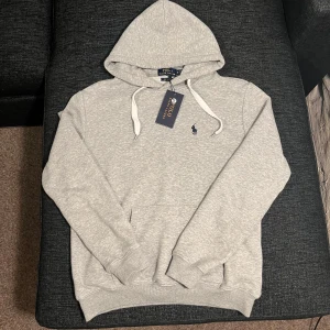 Grå hoodie från Ralph Lauren - Säljer en helt ny stilren grå hoodie från Ralph Lauren med klassisk passform. Den har en justerbar huva med vita snören och det ikoniska logotypbroderiet på bröstet. Perfekt för en avslappnad look. Gråa snören är inkluderade. Perfekt som julklapp till pojkvännen eller brorsan! Tveka inte på att höra av er vid funderingar. Mvh William