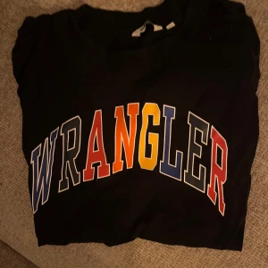 Svart t-shirt från Wrangler - Snygg svart t-shirt från Wrangler med färgglad text i blått, rött, gult och vitt. Perfekt för en casual look.