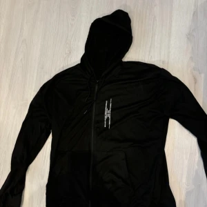 Svart huvtröja med dragkedja - Snygg svart huvtröja med dragkedja och textdetalj på framsidan. Tillverkad i bomull och polyester, perfekt för en casual look Eller träning. Huvan har justerbara snören för extra komfort.