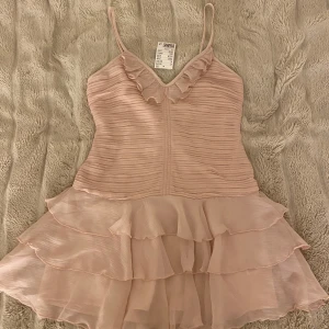 Ljusrosa klänning från H&M - Söt ljusrosa klänning från H&M med volangdetaljer och smala axelband. Klänningen har en omlott-design och flera lager av volanger som ger en fin figur. Köpte i somras men den är helt oanvänd 💕
