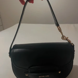 Svart axelväska från Michael Kors - Snygg svart axelväska från Michael Kors i skinn med gulddetaljer. Väskan har en justerbar axelrem och stängs med magnet. Perfekt för att bära dina nödvändigheter med stil. Använd enstaka ggr 
