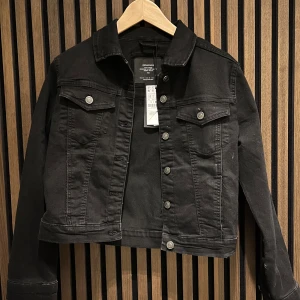 Svart jeansjacka från Stradivarius - Snygg svart jeansjacka från Stradivarius med klassisk design och knappar framtill. Jackan har två bröstfickor med lock och knappar, samt långa ärmar. Perfekt för en stilren look.