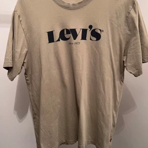 Beige t-shirt från Levi's - Säljer en beige t-shirt från Levi's med deras klassiska logga tryckt i svart på framsidan. T-shirten har en avslappnad passform och är tillverkad i mjuk bomull. Perfekt för en casual look.