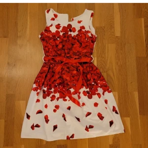 Vit och röd klänning med blommönster - Klänning med ett vackert blommönster på vit bakgrund. Klänningen har ett satinbälte och en dragkedja bak för enkel påklädning. Fin röd färg perfekt inför jul och nyår. Står ej storlek, men passar XS.