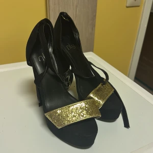 Svarta klackskor med glittrig detalj från Nly - Snygga svarta klackskor från Nly med en glittrig gulddetalj framtill. Skorna har en hög klack och en elegant design som passar perfekt för festliga tillfällen.