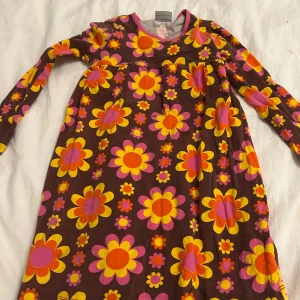 Blommig klänning från me&i - Härlig klänning från me&i med färgglatt blommönster i rosa, gult och orange på brun bakgrund. Klänningen har långa ärmar och en bekväm passform, perfekt för lekfulla dagar. Den kommer tvättas och strykas innan den skickas! ⭐️