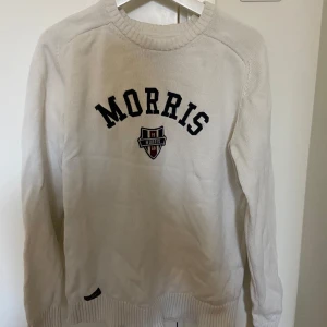 Vit stickad tröja från Morris  - Säljer en stilren vit tröja från Morris Gents med svart text på bröstet. Tröjan har långa ärmar och ribbade muddar vid ärmslut och nederkant. Perfekt för en avslappnad och snygg look.