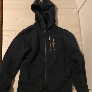 Mörkblå fleecejacka från Sail Racing - Säljer en mörkblå fleecejacka från Sail Racing med dragkedja och huva. Jackan har en broderad logotyp på bröstet och är perfekt för kyliga dagar. Den är långärmad och har en bekväm passform.