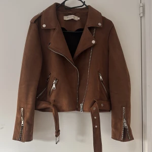 Höst jacka - Snygg brun mockajacka med dragkedjor och bälte från Senly Studio. Jackan har en klassisk bikerstil med långa ärmar och flera fickor med dragkedja i mjukt läder material. Jackan är i storlek L men passar Xs/S då den är liten i storlek. Bra skick inga defekter 