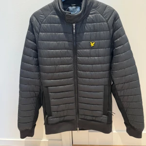Svart quiltad jacka från Lyle & Scott - Snygg svart quiltad jacka från Lyle & Scott med dragkedja och ribbade muddar. Jackan har en broderad logotyp på bröstet och en hög krage med knappar. Perfekt för kyligare dagar. Något nopprig vid de stickade partierna.