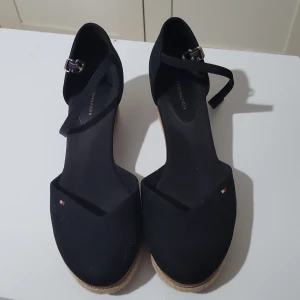 Svarta kilklackar från Tommy Hilfiger - Snygga svarta kilklackar från Tommy Hilfiger med justerbar rem runt ankeln. Skorna har en klassisk design med en liten logotypdetalj på framsidan och en bekväm kilklack i naturmaterial.