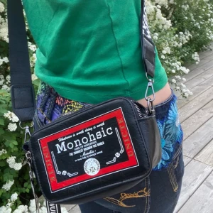 Svart axelväska med tryck - Cool svart axelväska med ett unikt tryck i rött och vitt. Väskan har en justerbar axelrem med text och en praktisk dragkedja för säker förvaring. Perfekt för att ge din outfit en edgy touch. Finns mycket utrymme och har plats med mindre saker som telefon, kort, kamera osv. 