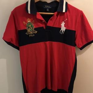 Röd och marinblå pikétröja från Ralph Lauren - Snygg röd och marinblå pikétröja från Ralph Lauren med broderad logga och nummerdetalj. Tröjan har en klassisk krage med knappar och korta ärmar. Perfekt för en stilren look. En stor barnstorlek så den är som S 